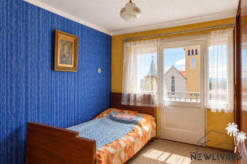 A room in a family house with a bed, blue wallpaper, and a view of the church.