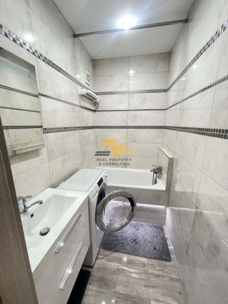 A bathroom in a studio apartment with a shower, a washing machine, and a wooden-patterned floor.