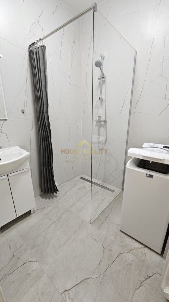 Bathroom with a shower in a 2-room apartment; sink, washing machine, light tiles.