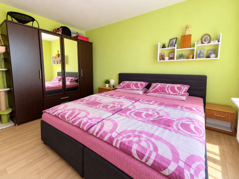 Bedroom in a 2-room apartment with a wood-patterned floor, green wall, and pink bedding.