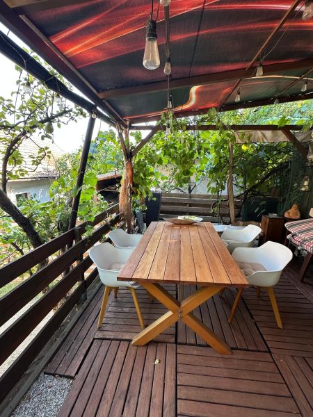 A terrace with a table and chairs under a pergola, surrounded by greenery and wooden boards.
