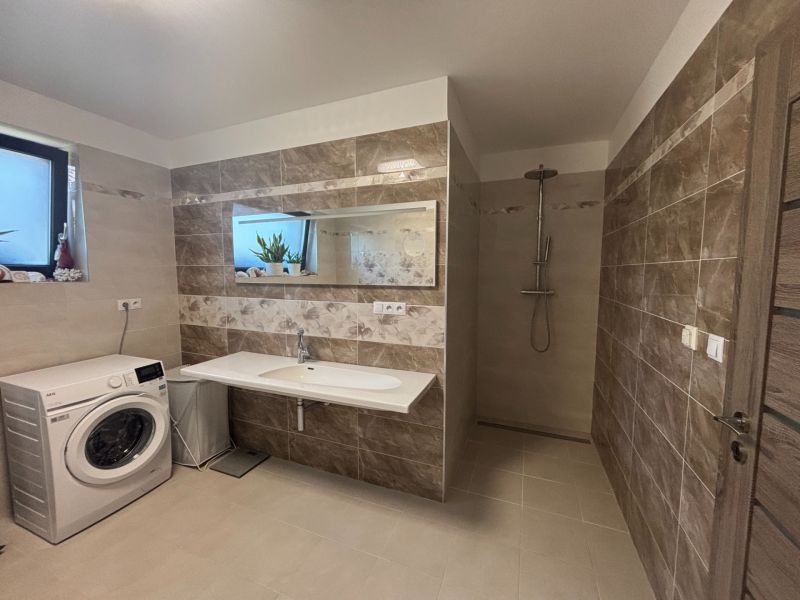 A bathroom in a family house with a washing machine, light tiles, and a shower corner.