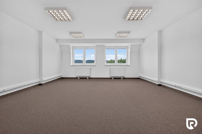 A spacious office with windows and carpeted flooring, lighting, and neutral walls.
