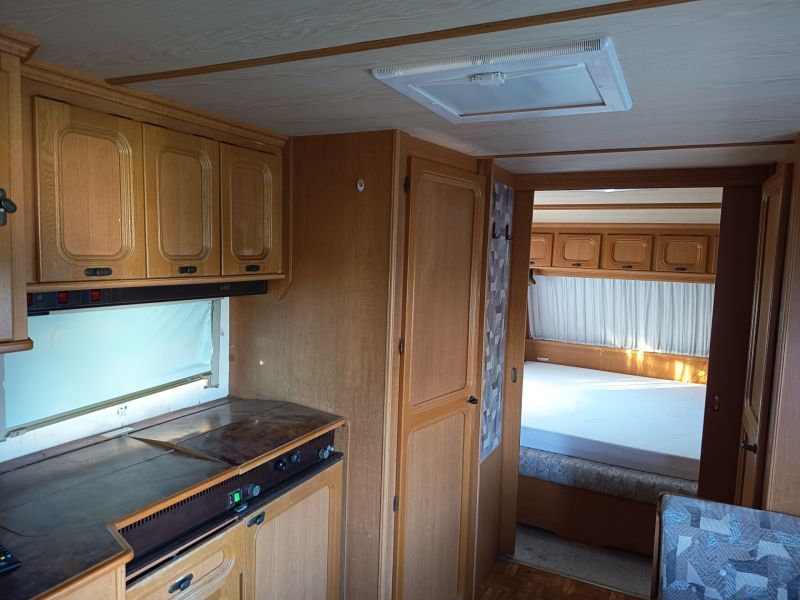Interior of a motorhome with a kitchenette and bedroom, wooden decor flooring, gardens.