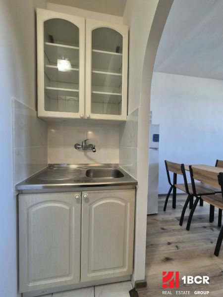 Kitchen corner in a studio apartment with a sink, cabinets, and a table on a wooden decor floor.