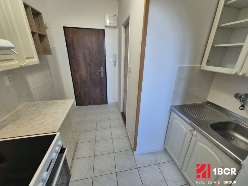 A kitchenette in a studio apartment with light cabinets and appliances, and a wood-patterned floor.
