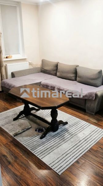 Living room of a three-room apartment with a sofa, table, wood-patterned floor, and carpet.
