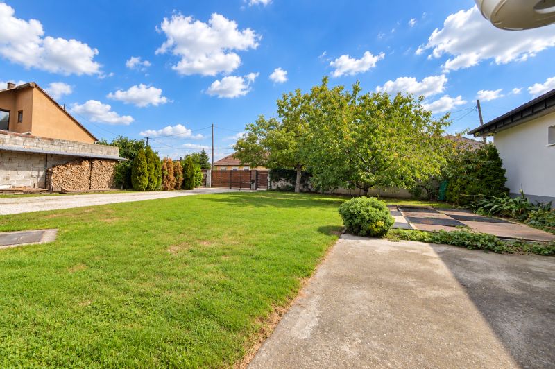 A garden in Mosonmagyaróvár with a lawn, trees, and a concrete path near a family house.