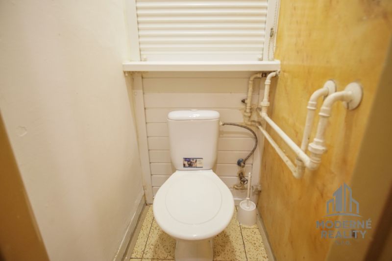 A small bathroom in a two-room apartment with a toilet, white walls, and pipes on the side wall.