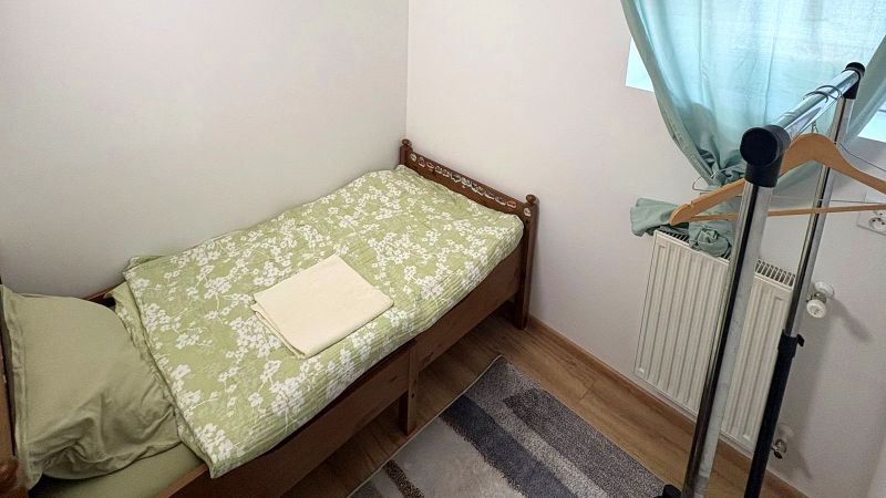Single bed, clothing rack, green curtains, wood-patterned floor.