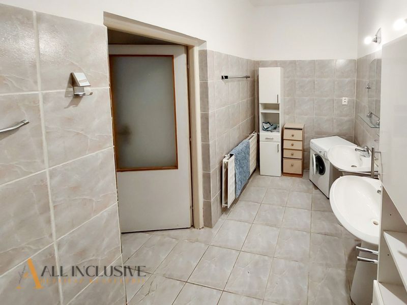 A bathroom in a two-room apartment with a washing machine, double sink, and cream-colored tiles.