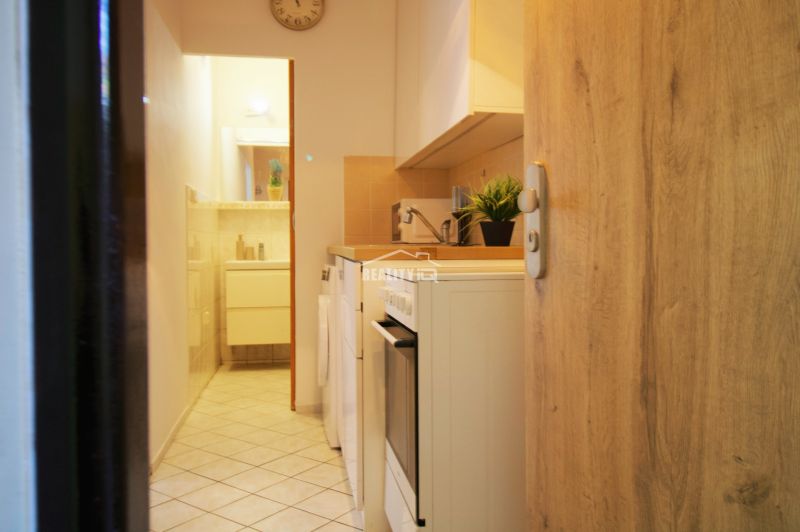 A kitchenette in a studio apartment with white cabinets and appliances, tiled flooring.
