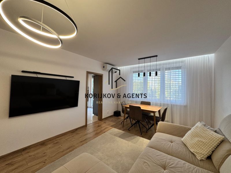 Living room in a 3-room apartment with a large television, wooden decor flooring, and a dining table.