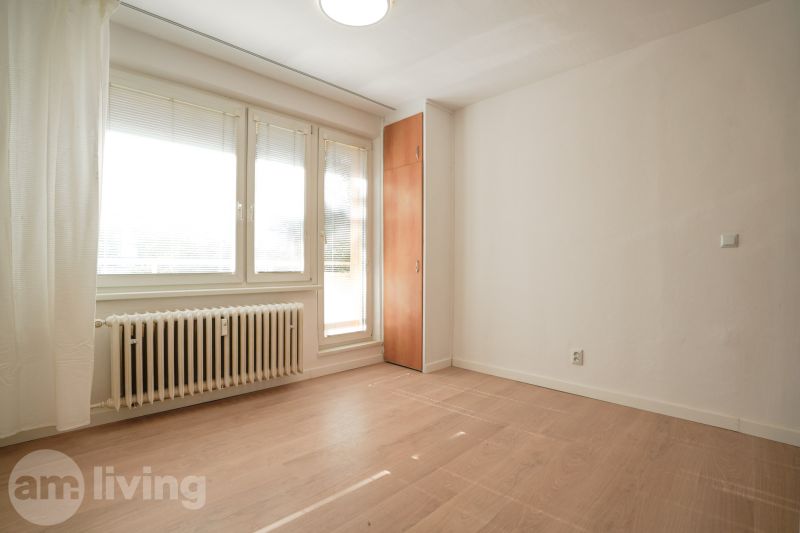 A room in a one-room apartment with a wooden decor floor, white walls, and windows.