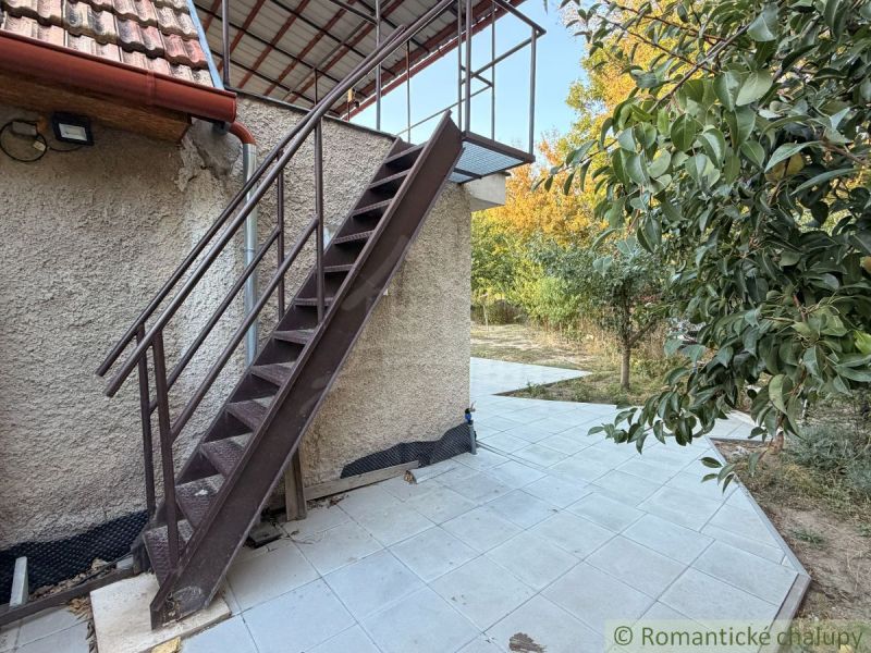 The exterior of a cottage in Nové Zámky with a metal staircase and a terrace surrounded by greenery.