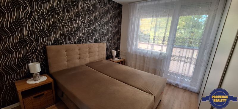 Bedroom with upholstered bed, patterned wallpaper, curtains, wooden decor flooring.