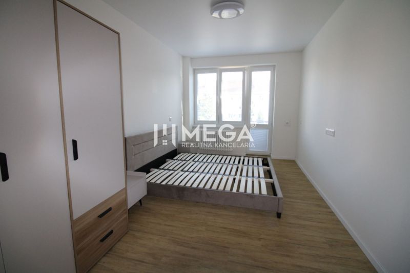 A bright bedroom in a two-room apartment with wood-patterned flooring and a large window.