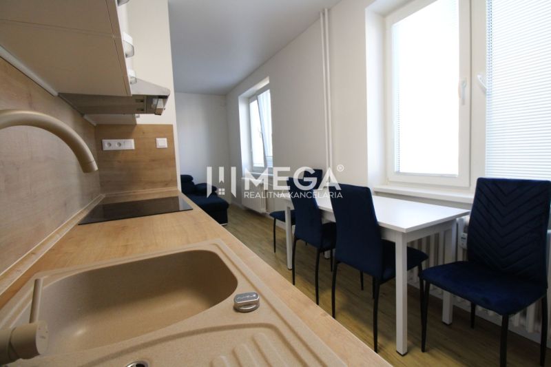 A kitchen in a 2-room apartment with white furniture, blue chairs, and a wooden-patterned floor.