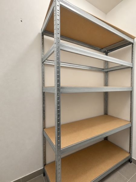 A metal shelf unit with shelves in a 2-room apartment on a white wall and grey floor.
