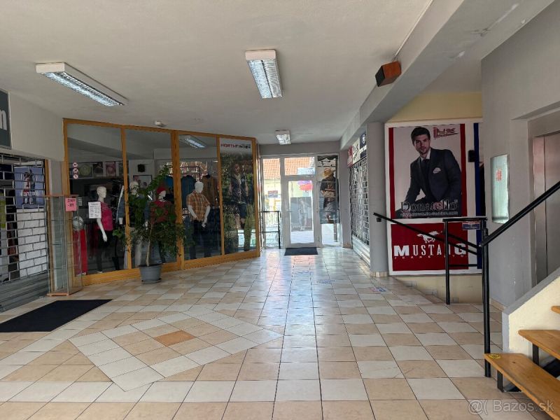 A corridor of commercial spaces with glass displays, a plant, and an advertisement on the wall.
