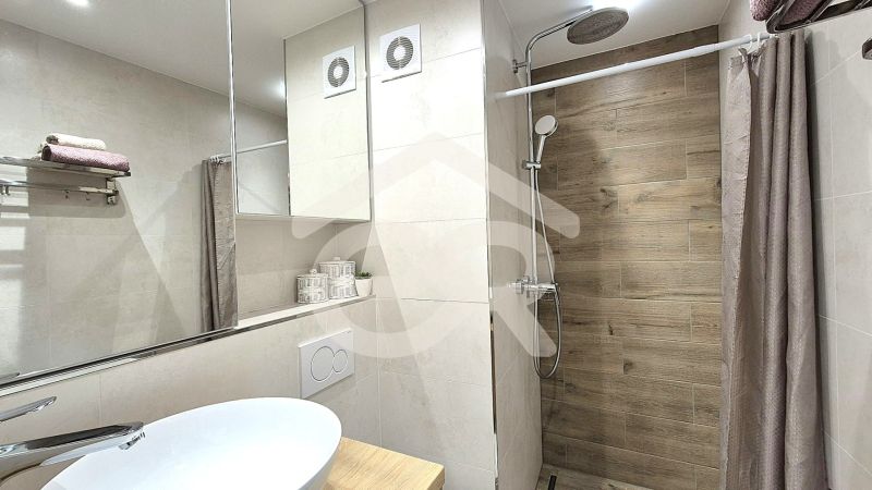 A studio apartment bathroom with a large mirror and a shower corner with a wooden decor.