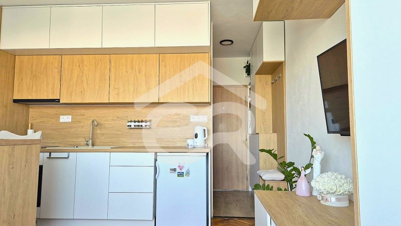 The kitchen part of a studio apartment with white and wooden cabinets, a television, and decorations.