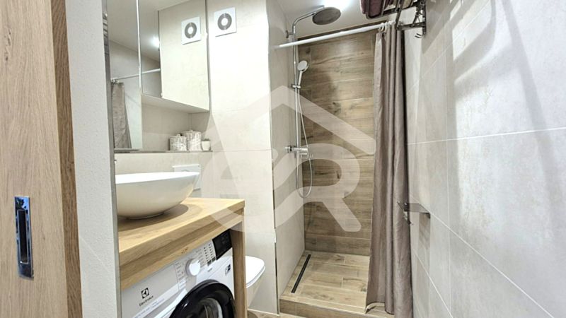 A bathroom in a studio apartment with a wooden decor floor, a shower, and a washing machine.