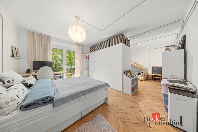 A bedroom in a two-room apartment with a wood-patterned floor, a large bed, and storage spaces.
