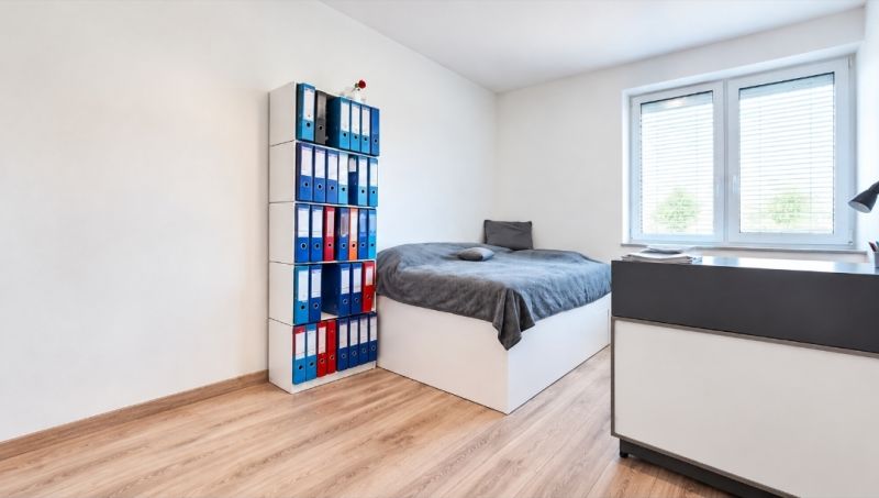 A bright room in a family house with a bed, a shelf for binders, and a floor with a wooden decor.