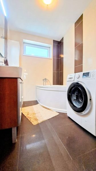 A bathroom in a family house with a bathtub, a washing machine, and dark tiles in a decorative design.